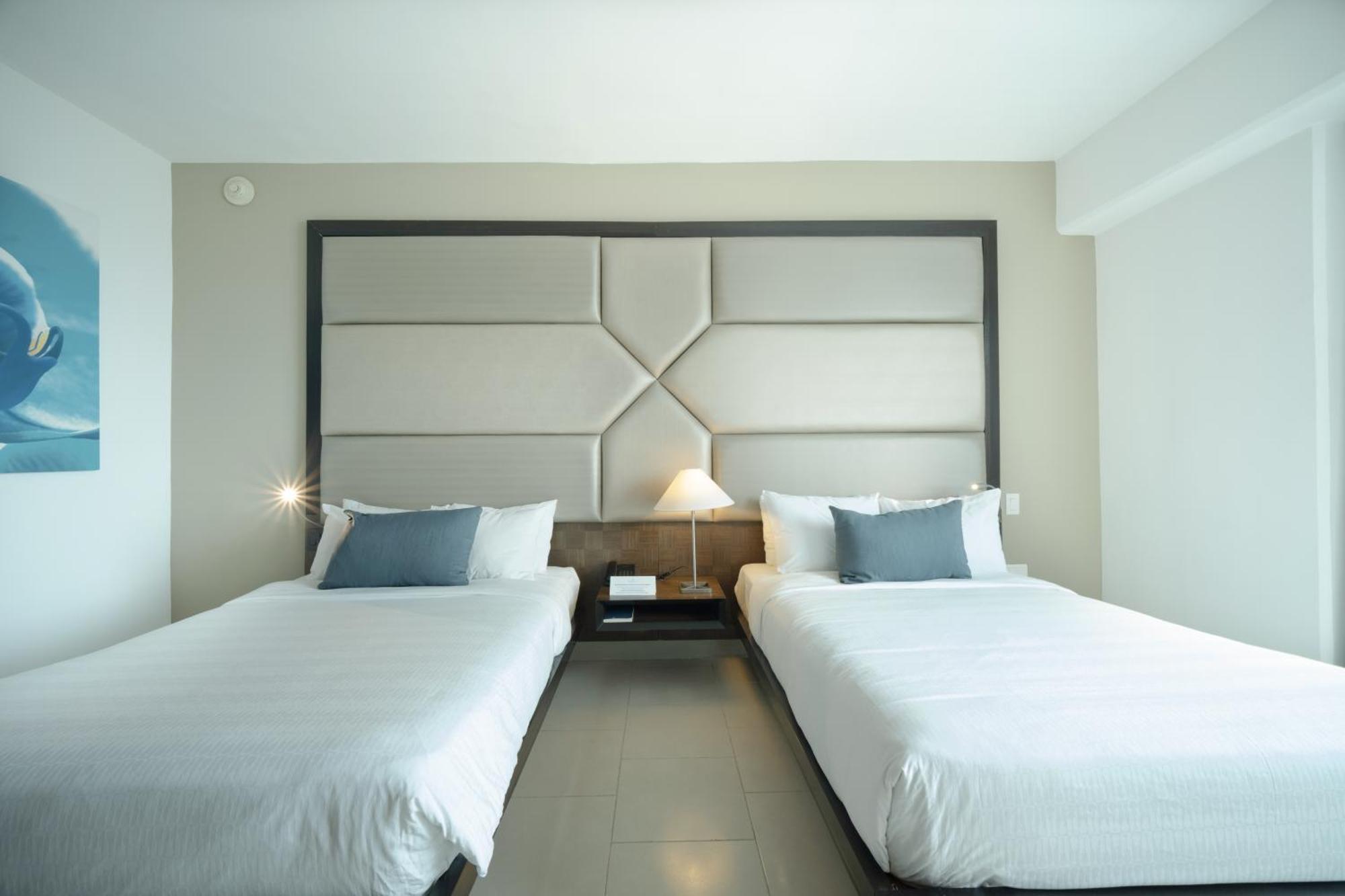 Decapolis Hotel Panama City Exterior photo The photo shows a modern hotel room with two neatly made beds, each with white bedding. The beds are positioned next to each other, with a contemporary headboard behind them featuring a geometric design. There is a small bedside table between the bed