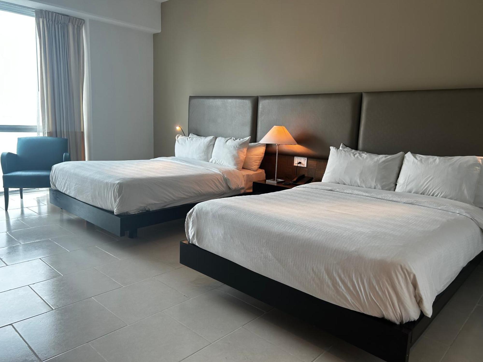 Decapolis Hotel Panama City Exterior photo The photo shows a hotel room featuring two large beds with white beddings. There is a bedside table with a lamp next to one of the beds. A cozy chair is placed in the corner of the room. Large windows allow natural light to fill the space, and the wa