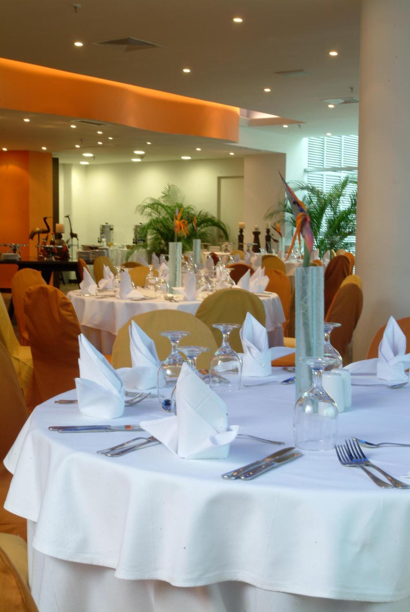 Decapolis Hotel Panama City Restaurant photo The photo shows a well-set dining area in a restaurant. There are elegantly arranged tables covered with white tablecloths, and each table features neatly folded napkins. Glassware, including wine glasses and water glasses, is placed on the tables. T