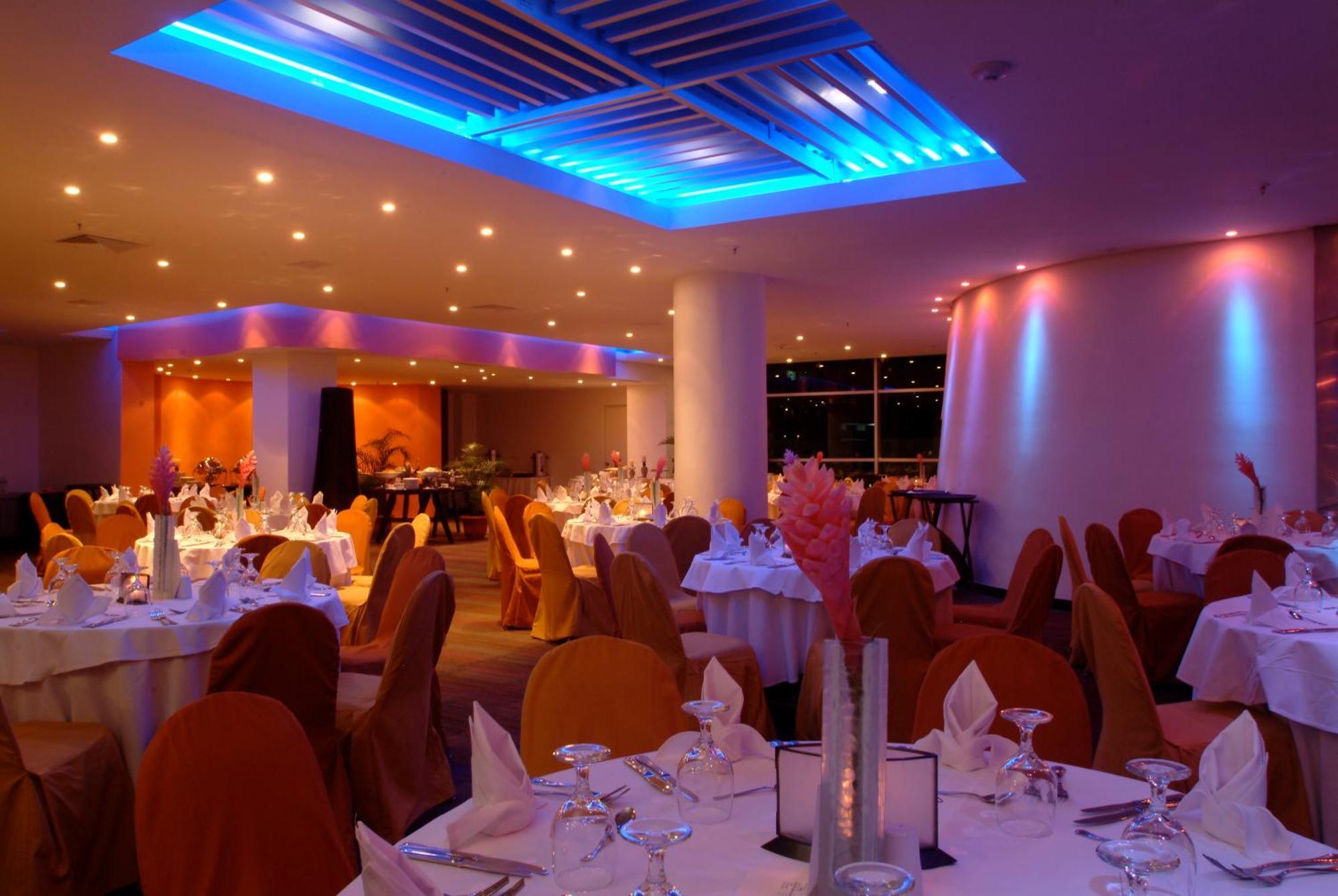 Decapolis Hotel Panama City Facilities photo The image shows an elegant restaurant interior. The dining area features round tables covered with white tablecloths, and each table is set with glassware and napkins. The chairs are draped in orange fabric, contributing to a warm and inviting atmosp